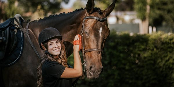 Smiling woman grooming a saddled horse outdoors on a sunny day.