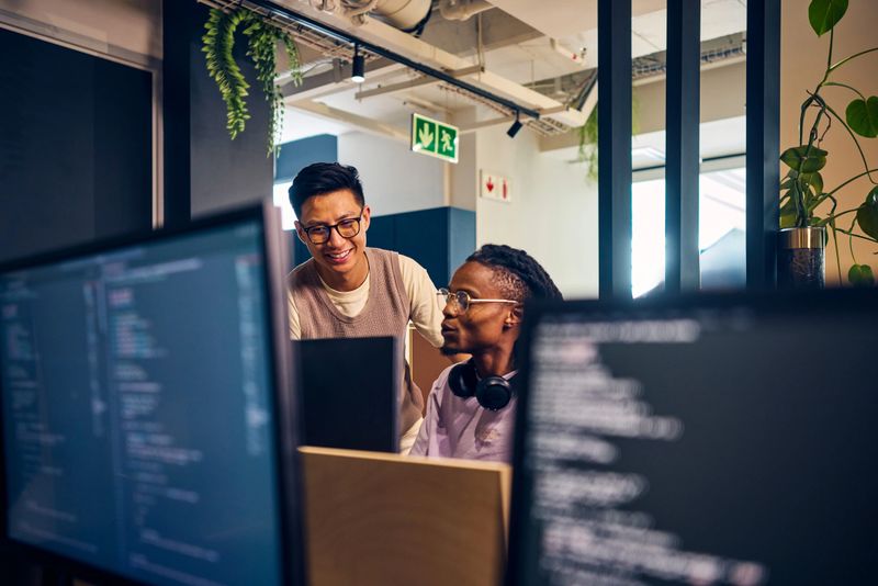 Two diverse software developers collaborating on a project, reviewing code on computer screens in a contemporary office space