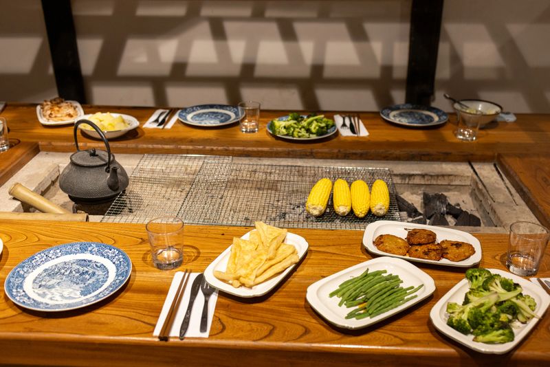 A beautifully set rustic dinner table featuring a variety of dishes, including grilled corn on the cob, sautéed broccoli, green beans, potato croquettes, and thinly fried tofu slices. A traditional cast-iron teapot adds a warm, homely touch, while blue-patterned ceramic plates enhance the timeless charm of the setting. The wooden table and ambient lighting create a cozy dining atmosphere, perfect for family gatherings or casual dinner parties.