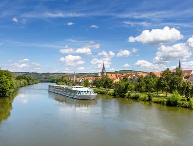 River cruise boat on a calm river with a town and cloudy blue sky.