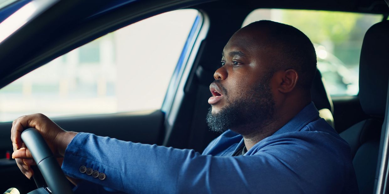 Man in blue jacket driving a car, looking surprised or shocked.
