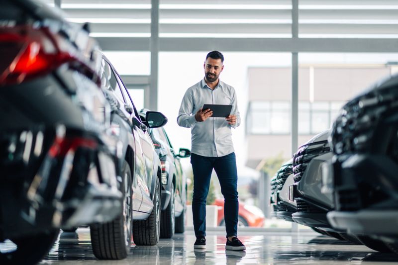 A man in a grey shirt is captured using a tablet to meticulously inspect a range of cars in a contemporary dealership's showcase area, highlighting modern sales techniques.