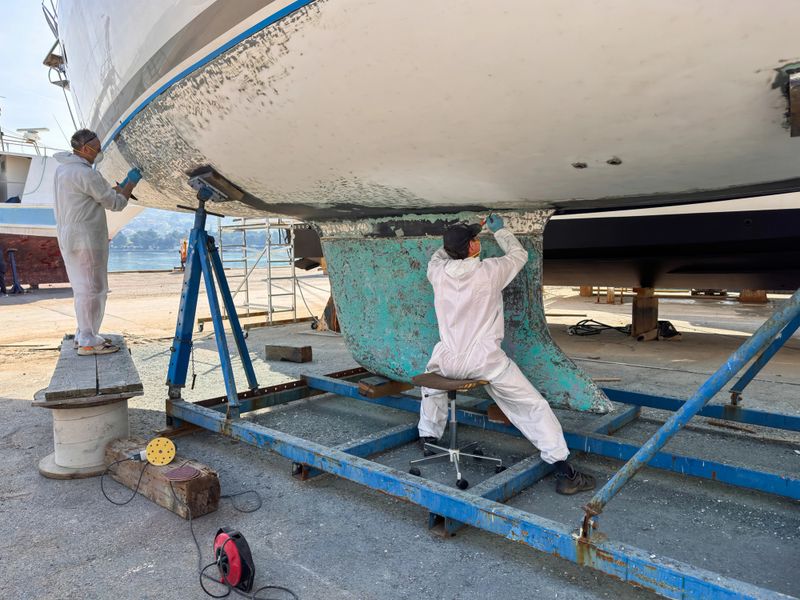 Men wearing protective gear removing antifouling paint with paint scraper from sailboat keel and hull.
