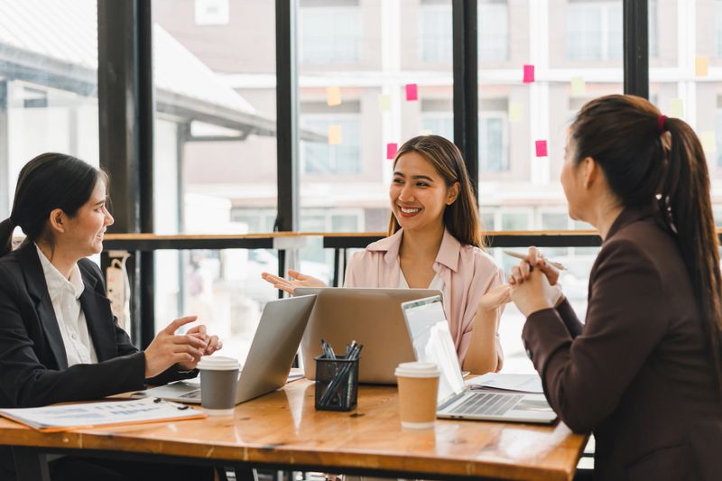 Efficient teamwork is showcased in this modern office setting, where three women engage in productive discussion around table filled with laptops and coffee cups. Their expressions reflect.
