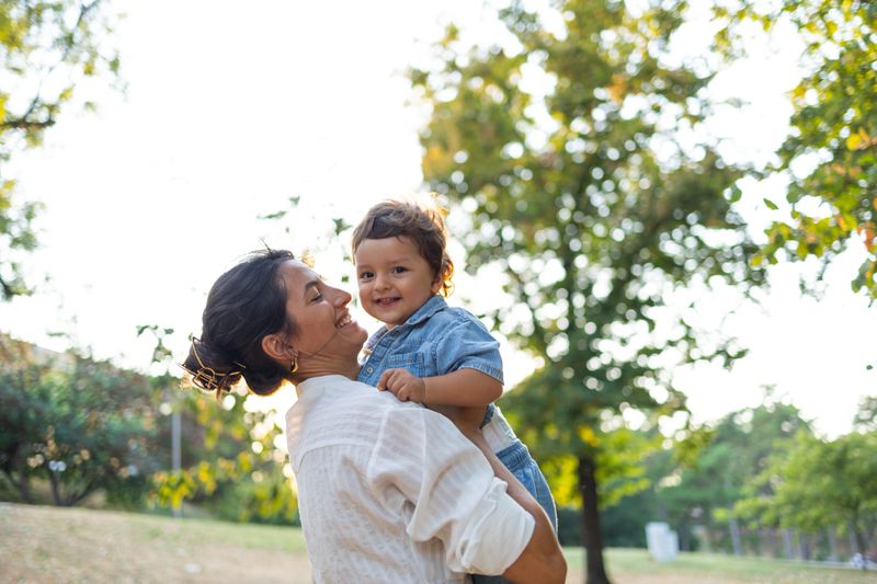 A smiling mother lifts her happy child in a sunny park, surrounded by lush green trees. The moment captures the joy and warmth of a family day outdoors.