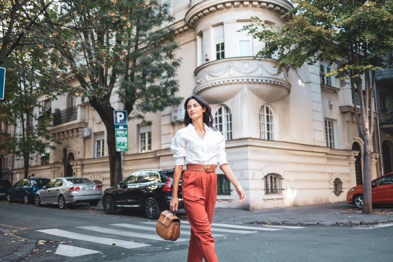 A stylish woman walks confidently down a picturesque street in a charming urban neighborhood, exuding elegance and freedom. She carries a woven handbag and is surrounded by classic architecture and parked cars.