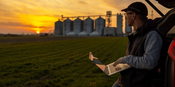 Homem com laptop observa o pôr do sol em campo agrícola.