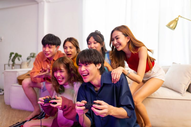 Group of Young Asian man and woman playing video games console together in living room at home. Happy generation z people friends enjoy and fun indoor lifestyle meeting party on holiday vacation.