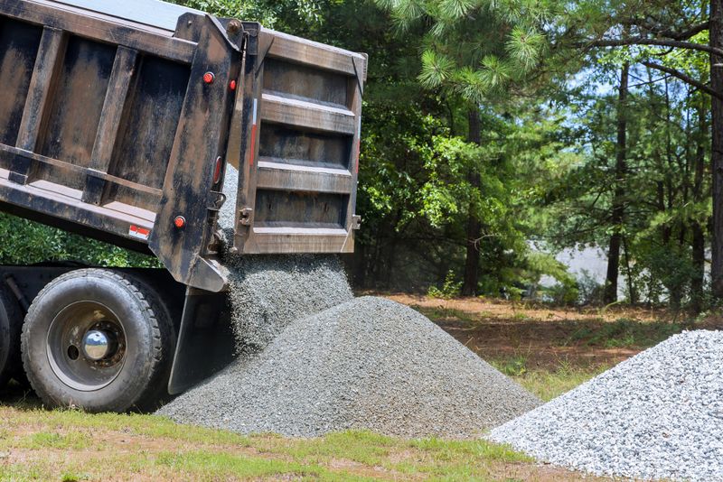 Truck unloads gravel onto grassy area preparing for landscaping work process demonstrates delivery of construction materials for improvement projects.