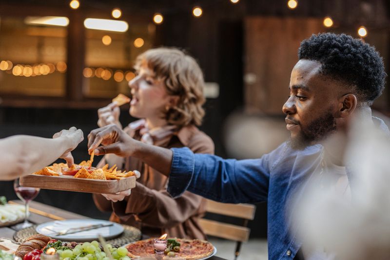 Group of multi ethnic friends enjoying a meal together outdoors, sharing nachos with salsa and other snacks like pizza and grapes during a casual celebration