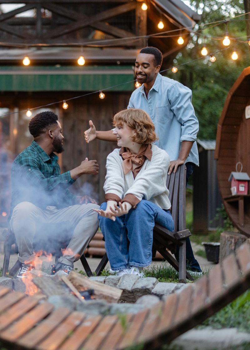 Three cheerful multiracial friends are sitting around a bonfire, chatting and enjoying a cozy outdoor party in the backyard of a wooden house