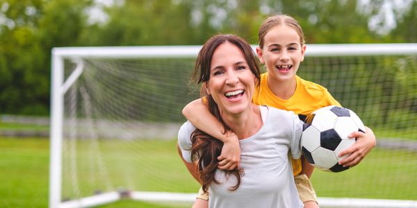 Smiling mother giving her daughter a piggyback with a soccer ball on a field.