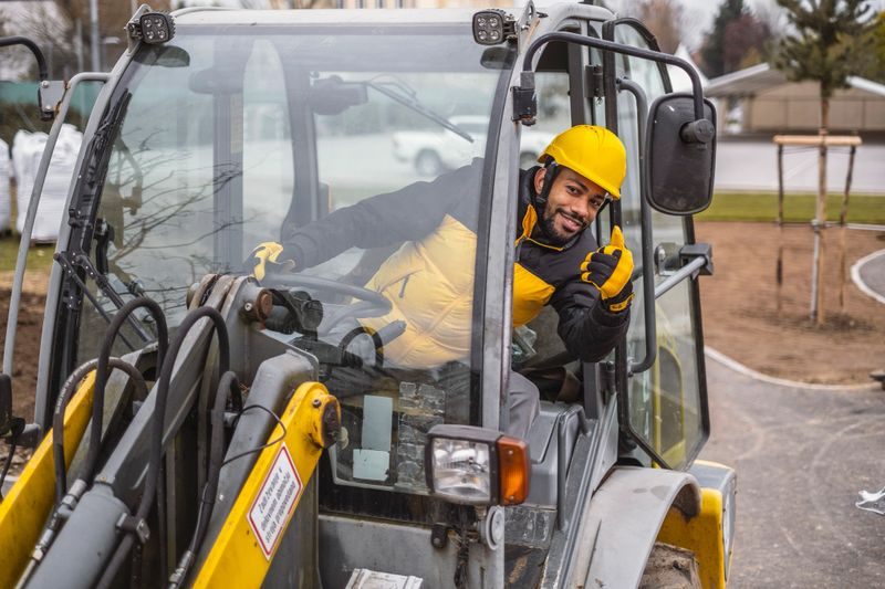 A mixed-race male construction professional sports a hard hat and high-visibility jacket while adeptly maneuvering a mini excavator in an outdoor setting, showcasing skillful earthmoving tasks.