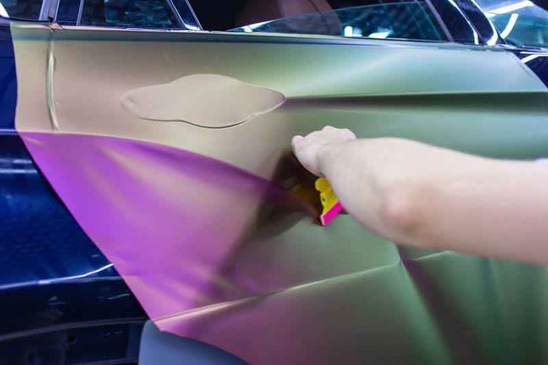 A specialist in wrapping a car with chameleon-colored vinyl film in the process of work. Car wrapping close-up. Car wrapping specialists cover the car with vinyl sheet or film. Selective focus.