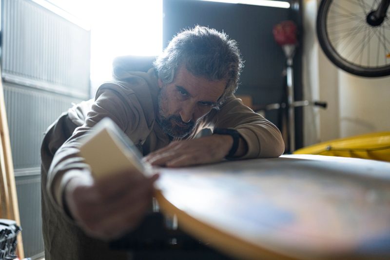 Green Business. Mature man repairing surfboard surface dings, searches for damage to the shell or core. Surfboard repair technician day in the life.