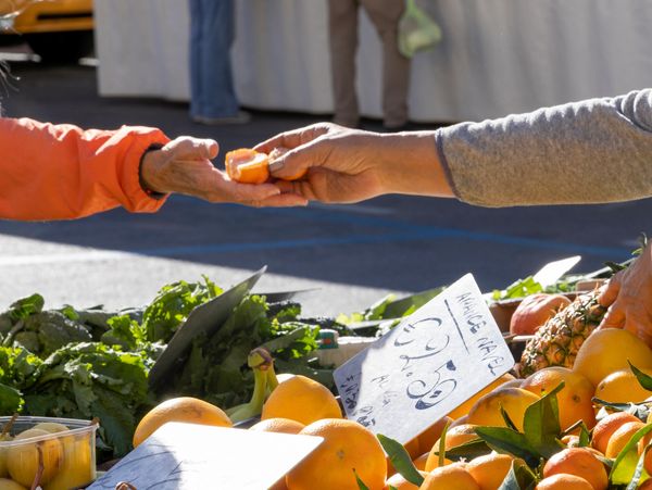 A vendor offering a customer a sample of an orange at an outdoor produce market.