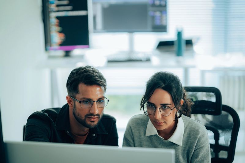 Two programmers collaborating in a modern workspace, analyzing code on-screen with reflections on glasses, symbolizing innovation and teamwork.
