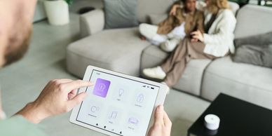 Person controlling smart home devices on a tablet with a couple in the background.