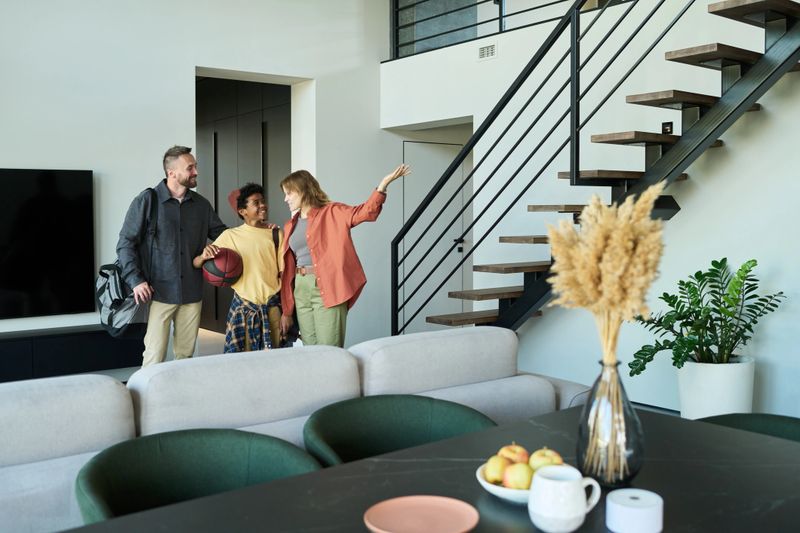 Happy family entering well-lit, modern home with minimalistic decor and wooden staircase. Basket of fresh fruit on table in foreground