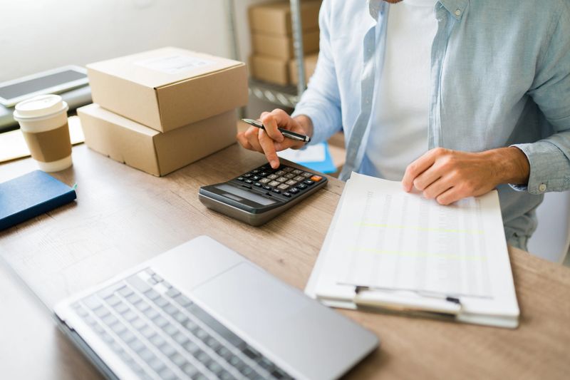 Small ecommerce business owner using calculator and reviewing logistics documents, managing orders and finances for online store