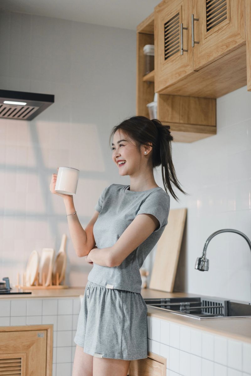 young woman in gray outfit enjoys cup of coffee in modern kitchen, radiating happiness and relaxation. warm wooden cabinets and bright space enhance cheerful mood.