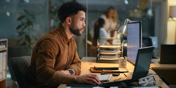 A focused man working late at his desk with computer screens in an office.