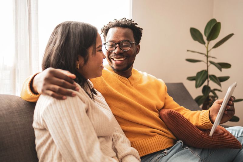 A joyful couple sits closely on a cozy sofa, sharing a moment while viewing a digital tablet. They display warmth and connection in a comfortable home setting.