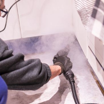 Person cleaning floor corners with a steam cleaner emitting steam.