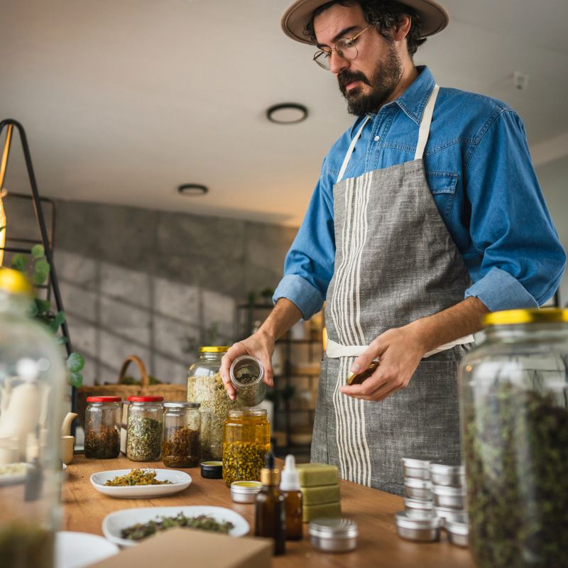 Side view of adult man herbalist hold jar with herbs an mix with oil in workshop