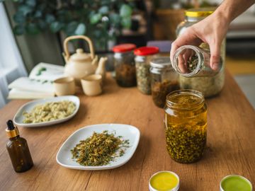 Hand pouring herbs into a jar on a wooden table with various jars and herbal items.