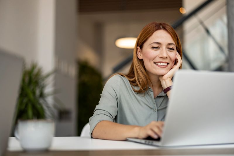 A smiling woman sits at a desk, working on her laptop in a contemporary, well-lit workspace. She appears engaged and content, suggesting a positive remote working experience.