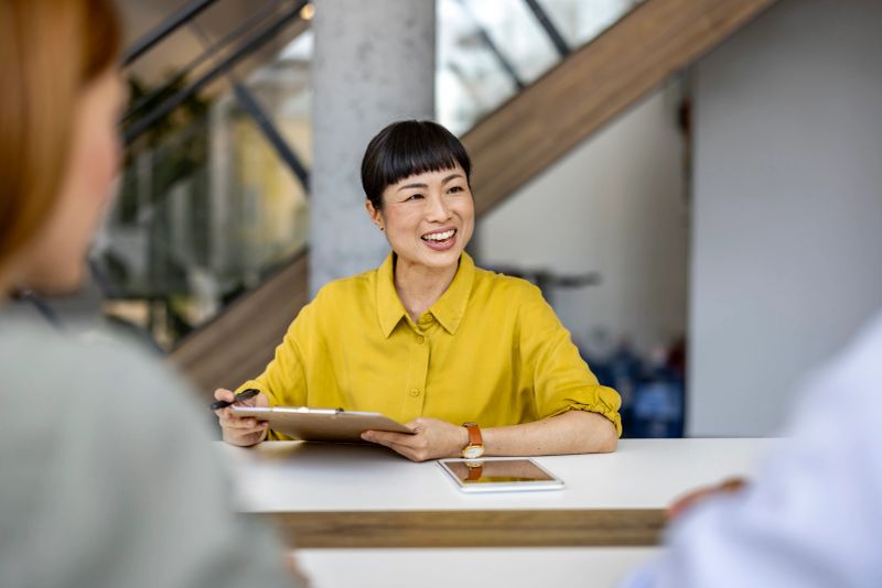 A smiling businesswoman sitting at a desk, leading a meeting and engaging with colleagues in a modern office setting. The atmosphere is positive and collaborative.
