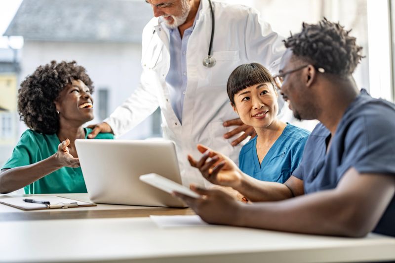 A diverse group of medical professionals in a discussion around a laptop in a bright office setting, showcasing teamwork, collaboration, and professional interaction in a healthcare environment.