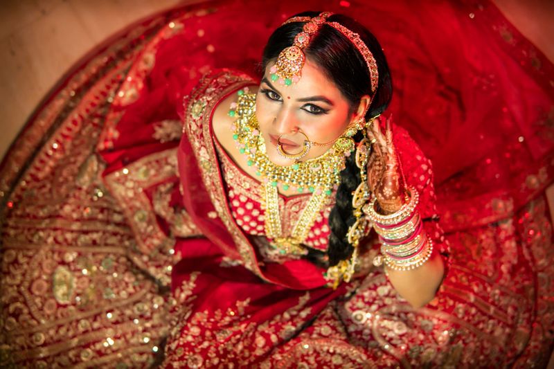 High angle portrait of beautiful, Asian, Indian bride wearing traditional bridal lehenga with heavy gold jewelry on her wedding day. She is looking up at the camera and smiling.