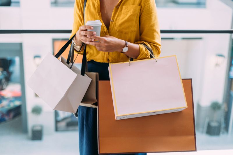 A stylish woman in a yellow shirt carries multiple shopping bags and a coffee cup in a modern mall setting, symbolizing consumerism and leisure.