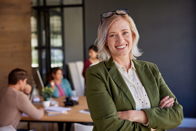 Portrait of confident mid adult businesswoman standing with a smile in modern office. Professional business woman in an creative modern office environment, while supportive team working in the background. Empowered executive woman, embodying strength and positivity in the workplace.