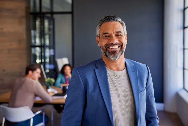 Smiling happy business man standing confidently in modern office. Proud indian entrepreneur standing in office with team working in background. Successful mid adult businessman looking at camera with supportive business team in background.