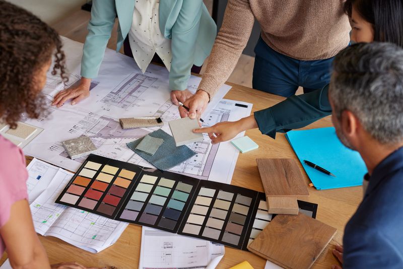 Top view of multiethnic people selecting materials and colors for a new housing project. Interior designers discussing color palette and material samples. High angle view of group of mixed race businesspeople reviewing plans with samples on the table.