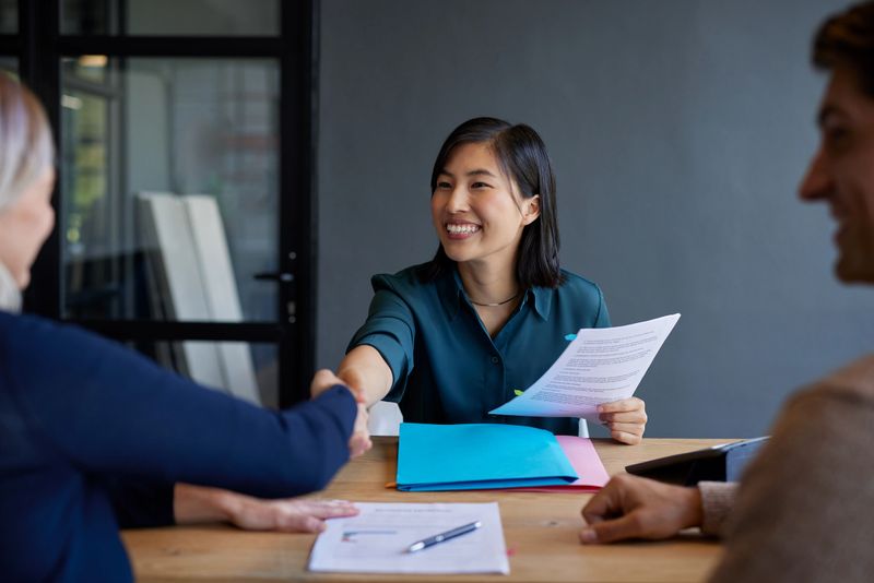 Smiling chinese candidate shakes hands during a job interview with female boss. Professional interview with candidate presenting documents. Financial advisor shaking hands after meeting a couple interested in new investments.