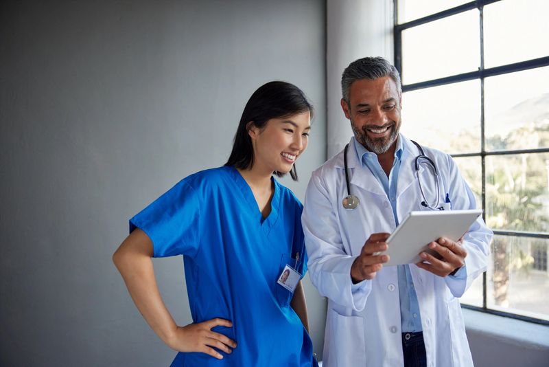 Happy mid adult doctor and a chinese nurse smiling while reviewing information on digital tablet. Medical professionals collaborating with positive energy at modern hospital. Healthcare workers discussing patient care in a bright office with copy space.