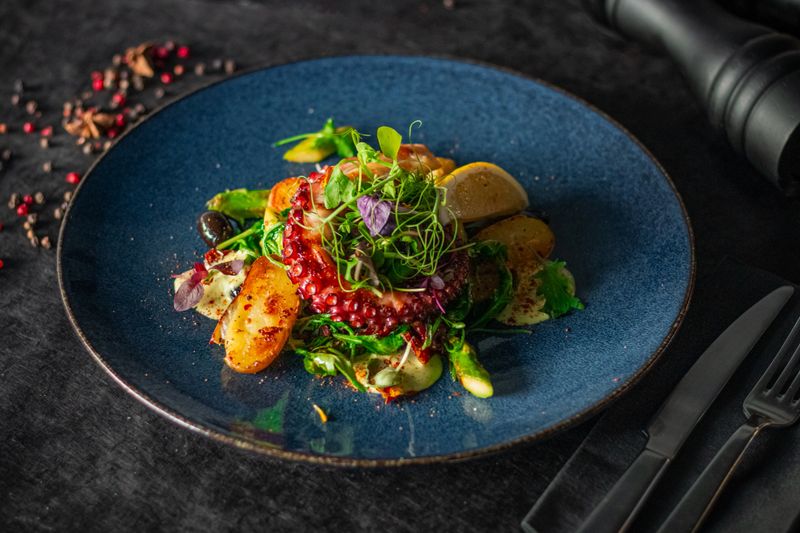 A vibrant octopus dish served with roasted potatoes, greens, and lemon slices on a blue plate, styled elegantly against a dark background with scattered spices.