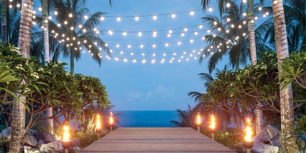 Wooden path leading to ocean with hanging lights and tiki torches.
