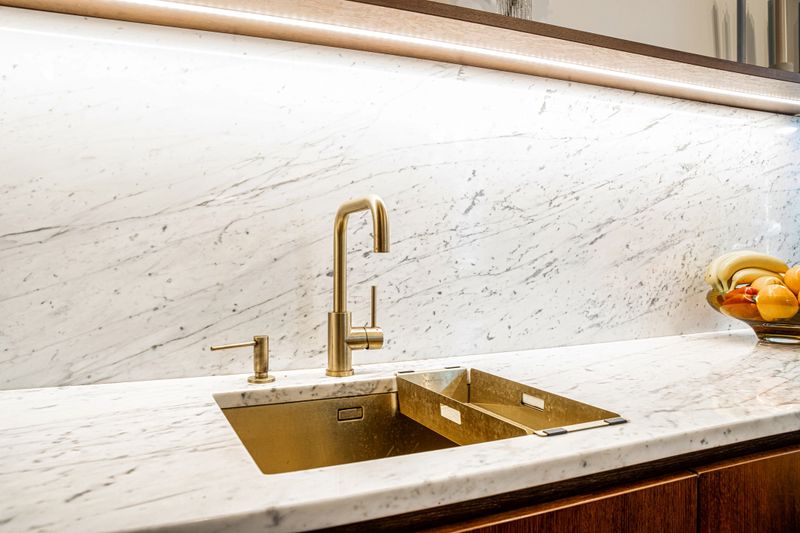 A luxurious kitchen features a marble countertop and backsplash, gold faucet and sink, and a bowl of fresh fruit, with under cabinet lighting.