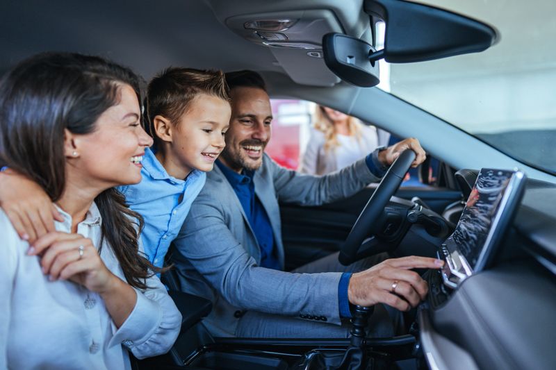 Excited family of three sits in a car at a dealership, exploring the vehicle's interior features. The father is demonstrating dashboard functions while the mother and son look on happily.