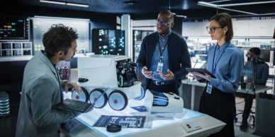 Three professionals discussing a robotic arm prototype in a lab.