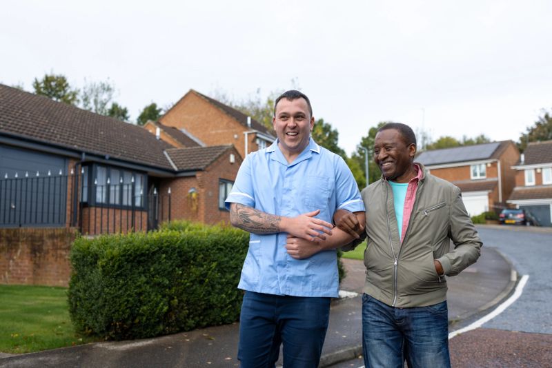 Medium three-quarter-length shot of a male community nurse walking with a patient in a residential area in Jarrow, North East England. They are laughing and talking as the patient interlinks one arm through the nurse's for support.

Videos similar to this scenario available.