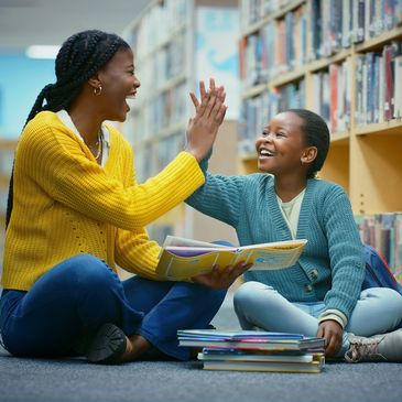 Two girls in a library high-fiving while sitting on the floor with books.