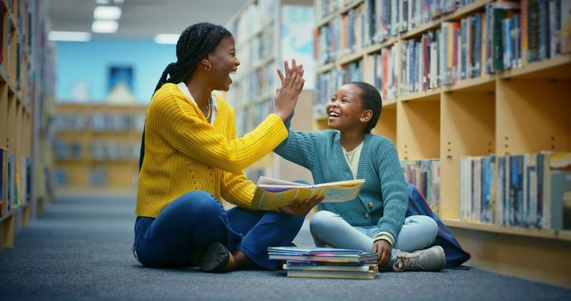 Library, girl and teacher high five for education goal, learning and celebration for storytelling. Educator, black woman and kid on floor for fun, fantasy stories and language development or relaxing