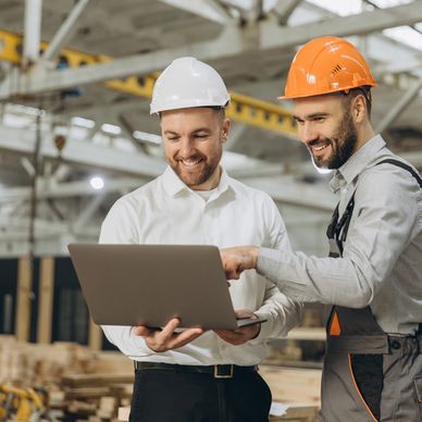 Two construction workers discussing plans on a laptop in a workshop.