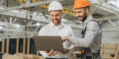 Two construction workers in helmets discussing a project on a laptop.